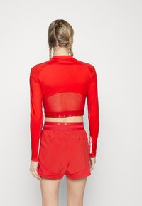 Czerwony krótki top z długim rękawem, z przezroczystym tyłem i błyszczącymi akcentami, zestawiony z czerwonymi szortami o luźnym kroju i elastycznym paskiem w talii.