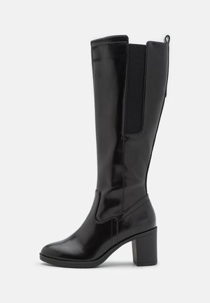 Botte noire montante en matériau synthétique brillant, dotée d'un talon bloc, de panneaux latéraux élastiques, et d'une pointe arrondie.