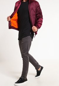 Bordowa kurtka bomberka z pomarańczową podszewką, czarny t-shirt, szare spodnie i czarne wsuwane buty. Kurtka ma prążkowany kołnierz i małe logo.