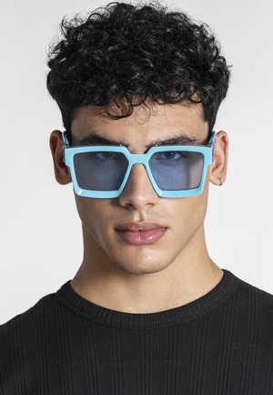 Man met krullend haar die grote, lichtblauwe vierkante zonnebrillen draagt en een zwart geribd shirt met ronde hals, tegen een effen achtergrond.