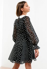 Mädchen trägt ein schwarzes, transparentes Kleid mit Punkten, langen puffigen Ärmeln und einem weißen Spitzenkragen, stehend vor einem weißen Hintergrund.