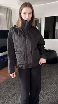Black quilted jacket with a high collar, featuring two front pockets and textured pattern. Worn over black pants, displayed indoors.