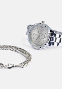 Silver watch with a round, stone-studded bezel and a metal link band; features a light-colored dial with numeric and decorative accents. Silver chain bracelet nearby.