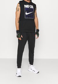 Sweat à capuche noir sans manches avec logo Nike blanc, joggers noirs et baskets blanches. Gants avec accents verts. Coupe ajustée, look sportswear décontracté.