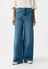 Široke denim hlače, svetlo-modre, visoko pasu, s petimi žepi in gumbom za zapiranje. Mehka tekstura z rahlo bledim videzom.