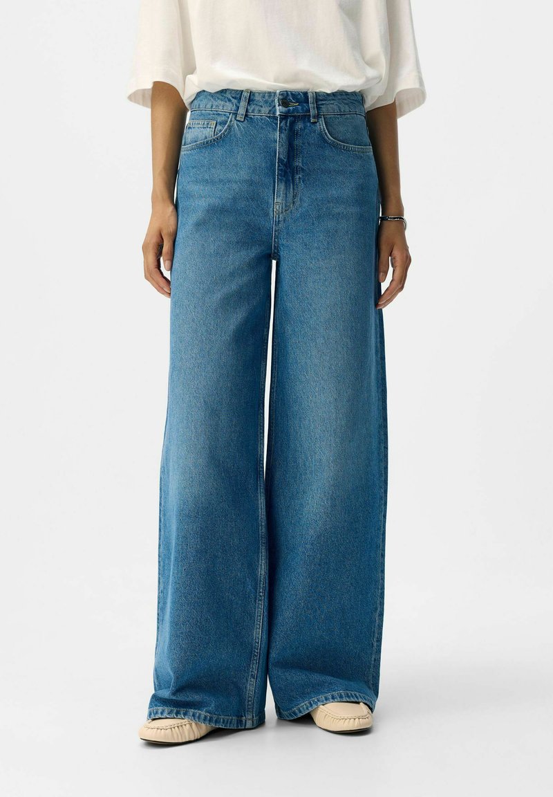 Široke denim hlače, svetlo-modre, visoko pasu, s petimi žepi in gumbom za zapiranje. Mehka tekstura z rahlo bledim videzom.