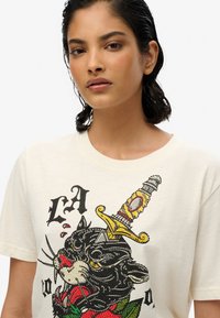 T-shirt en coton couleur crème mettant en avant un graphisme d'un chat noir avec une épée, agrémenté de roses rouges et d'accents décoratifs.