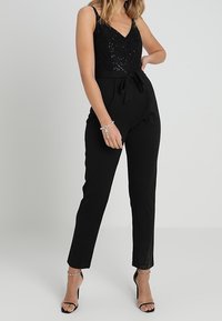 Mono negro con un cuerpo de lentejuelas, escote en V y un cinturón ancho como acento. Piernas ajustadas y tacones con correa en el tobillo completan el look.