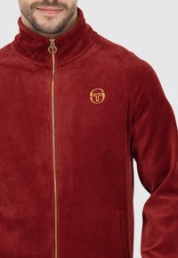 Rote Fleecejacke mit hohem Kragen, goldfarbener Reißverschluss und gesticktem Logo. Weiche Textur mit glattem Finish. Mit seitlichen Taschen.