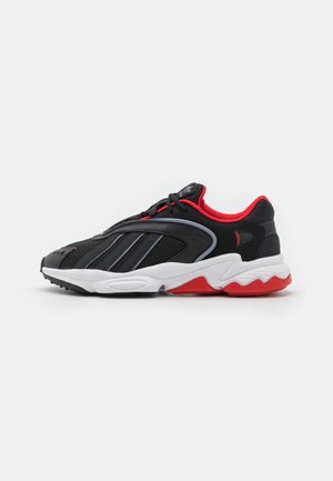 Schwarzer Sportschuh mit rotem Innenfutter, grauen Streifen, weißer Zwischensohle und roter sowie schwarzer Außensohle, seitlich auf weißem Hintergrund dargestellt.