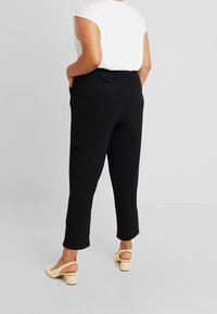 Osoba stojí zády k fotoaparátu, má na sobě bílé tričko a černé kalhoty, doplněné žlutými lodičkami.