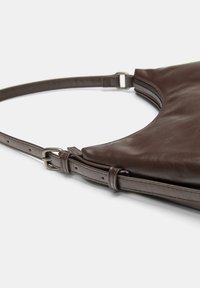 Bruine leren schoudertas met een gebogen ontwerp, verstelbare schouderriem en zilverkleurige hardware; heeft een ritssluiting aan de bovenrand.