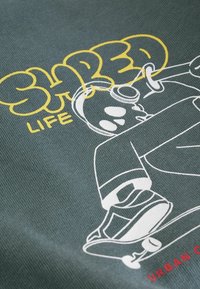 Tricou grafic cu un material verde închis, având un design alb de skateboard și text galben "SHRED LIFE" scris într-un font jucăuș.
