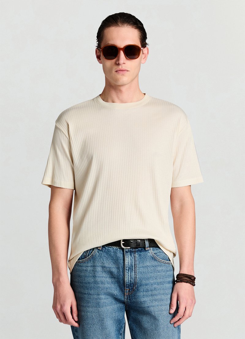 Beige geripptes T-Shirt mit kurzen Ärmeln, lockere Passform, kombiniert mit blauen Jeans. Das Modell trägt braune Sonnenbrillen und einen schwarzen, strukturierten Gürtel.