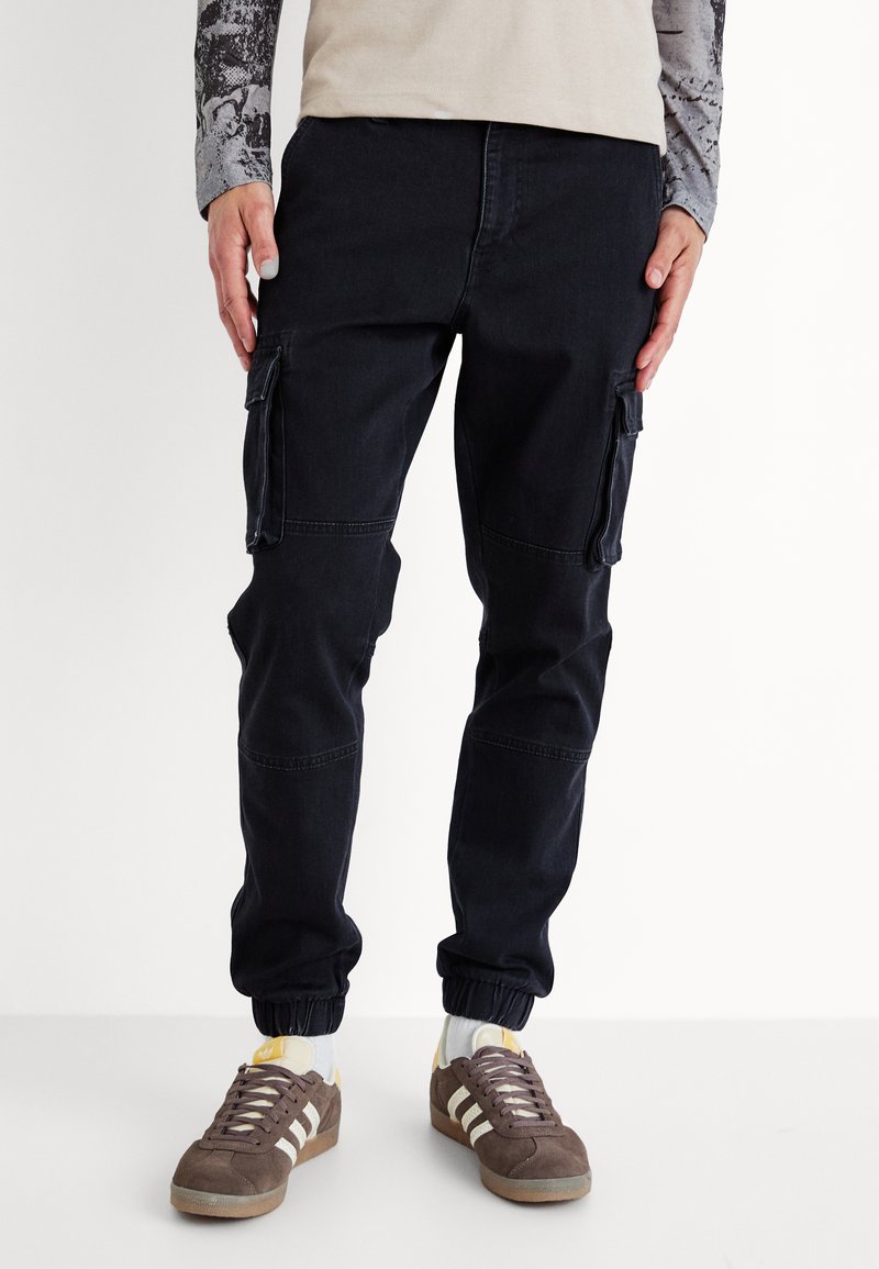 Pantalones cargo negros con puños elásticos, que cuentan con bolsillos laterales y un diseño ajustado. Llevados con zapatillas marrones y detalles en blanco.