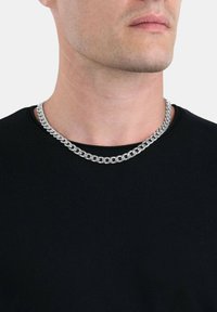 Zilveren ketting met in elkaar grijpende schakels, gedragen over een zwart ronde hals T-shirt, met een gladde, reflecterende oppervlakte en stevige uitstraling.