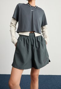 Šedý zkrácený sportovní top s krátkými rukávy, přes který je vrstvená texturovaná krémová dlouhá košile. S kombinací s tmavozelenými šortkami s elastickým pasem.