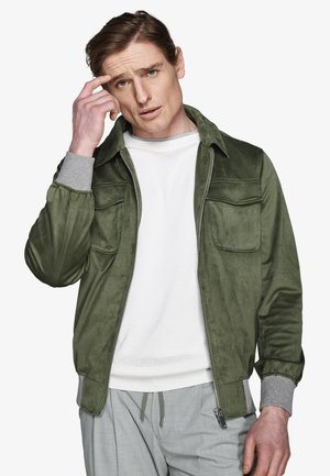 Mężczyzna w zielonej zamszowej kurtce, białej bluzie i szarych spodniach, dotykający skroni z zamyśloną miną na jednolitym białym tle.