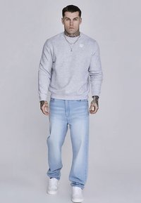 Ljusgrå sweatshirt med ribbstickade ärmslut, kombinerad med ljusblå jeans. Modellen bär vita sneakers och visar upp en avslappnad och ledig stil.