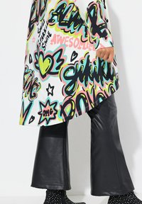 Tunique blanche avec un imprimé graphique multicolore vif, associée à un pantalon en cuir noir évasé et des bottines à pois.