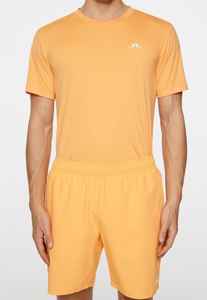 Hombre con camiseta de manga corta naranja a juego y pantalones cortos con pequeños logos blancos en el pecho y la pierna izquierda, de pie frente a un fondo blanco.