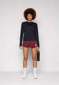 Čierne tričko s dlhým rukávom Nike, bordové šortky s bielym logom, biele ponožky a tenisky. Drží čiernu fľašu na vodu. Hladká textúra látky.