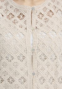 Close-up of cream crochet lace top with diamond patterns and four small white buttons down the center opening.