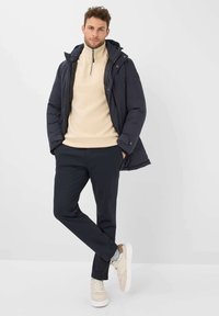 Giacca piumino blu navy, felpa bisaccia beige con mezza zip, pantaloni neri e sneakers beige. Dettagli notevoli includono cappuccio, accenti con zip e texture variegate.
