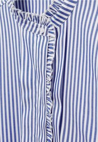 Gros plan sur une chemise à rayures verticales bleues et blanches avec un bord volanté le long de la patte de boutonnage et du col, présentant un unique bouton blanc.