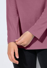 Dlouhý rukáv, světlé mauve tričko s kulatým výstřihem. Tkanina je jemná s mírným leskem. Ruka upravuje manžetu a odhaluje těsný střih.