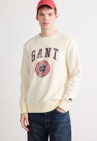 Kremowa bluza z logo "GANT", okrągłą naszywką graficzną, �żebrowanym dekoltem i mankietami. Połączona z niebieskimi dżinsami. Casualowy krój.