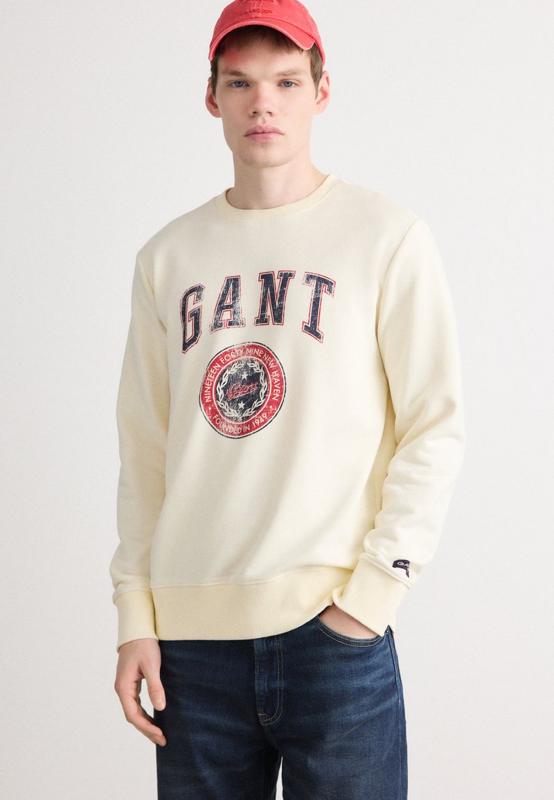 Kremowa bluza z logo "GANT", okrągłą naszywką graficzną, żebrowanym dekoltem i mankietami. Połączona z niebieskimi dżinsami. Casualowy krój.