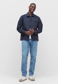 Marinblå bomullsjacka med knappframsidan, två bröstfickor och klassisk krage, kombinerad med ljusblå jeans och vita sneakers.