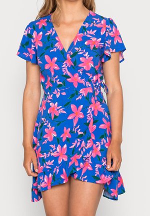 Niebieska sukienka kopertowa z różowym kwiatowym wzorem, krótkimi falbanistymi rękawami i falbanką u dołu, noszona przez osobę stojącą z rozluźnionymi ramionami.