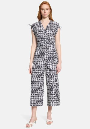 Kvinde iført en sort-hvid mønstret ærmeløs buksedragt med vidbenede ben, et bundet taljebælte og hvide åben-tå hælede sandaler.