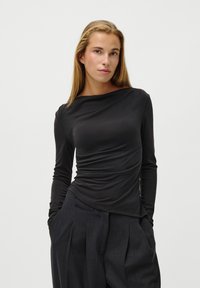 Crni dugi rukav s drapiranim dizajnom, širokim izrezom i mekanom teksturom. Nosite ga s labavim tamno sivim hlačama.