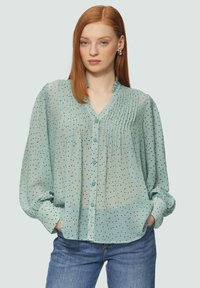 Jasnozielona, przezroczysta bluzka z dekoltem w kształcie litery V, plisowanym detalem i czarnymi kropkami. Ma długie rękawy i guziki z przodu.