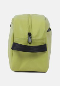 Rechteckige hellgrüne Tasche mit schwarzem Reißverschluss und schwarzem horizontalem Griff auf der Vorderseite, von der Seite vor weißem Hintergrund gezeigt.