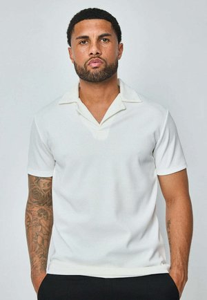 Polo à manches courtes en tissu texturé blanc, avec un col à revers et une coupe ajustée. Un style épuré et minimaliste avec des détails subtils.