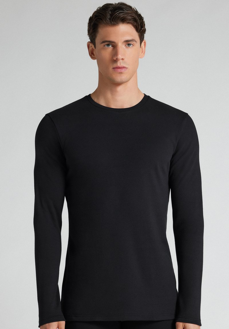 Jeune homme aux cheveux bruns courts portant un t-shirt noir uni à manches longues, debout devant un fond gris clair.