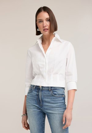 Frau trägt ein eng anliegendes weißes Hemd mit Dreiviertelärmeln und blaue hoch taillierte Jeans, steht vor einem hellen Hintergrund.