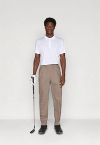 Man med vit polotröja, beige byxor, svarta golfsko och en handske, som håller en golfklubba och står mot en neutral bakgrund.