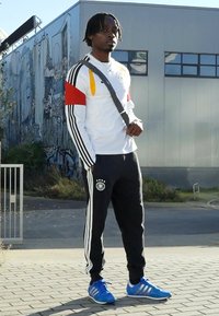 White long-sleeve shirt with red and yellow accents, black sweatpants with white stripes, blue athletic shoes, and a crossbody bag.