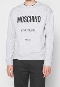 Šedá mikina s kulatým výstřihem, s výrazným černým nápisem "MOSCHINO COUTURE!" a "Milano" na přední straně. Měkká textura látky.