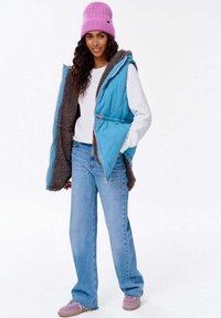 Modrý prošívaný vest, s měkkým hnědým interiérem, růžovými akcenty a bočními kapsami, nošený přes bílý svetr a spárovaný se světle modrými džíny.