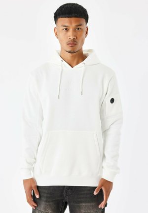 Sweatshirt à capuche blanche avec une poche avant, présentant un petit logo et un écusson texturé sur la manche. Le tissu est lisse avec une coupe décontractée.