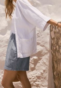 Femme portant une chemise blanche ample et une jupe bleue à fines rayures, debout avec un bras reposant sur un mur en pierre en plein soleil.