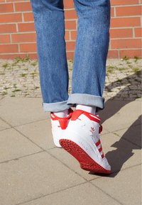 Sneaker bianche con accenti rossi, caratterizzate da un design alto, cinturino in Velcro e suola in gomma testurizzata con grip a motivo.