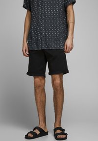 Svarta shorts med upplagda ärmar i kombination med en mönstrad kortärmad skjorta som har diamantformer i marinblått och vitt, kompletterat med svarta sandaler.
