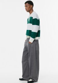 Grün-weiß gestreifter Pullover mit Kragen, kombiniert mit lockeren grauen Hosen. Der Pullover hat gerippte Bündchen; die Hosen haben große Taschen.